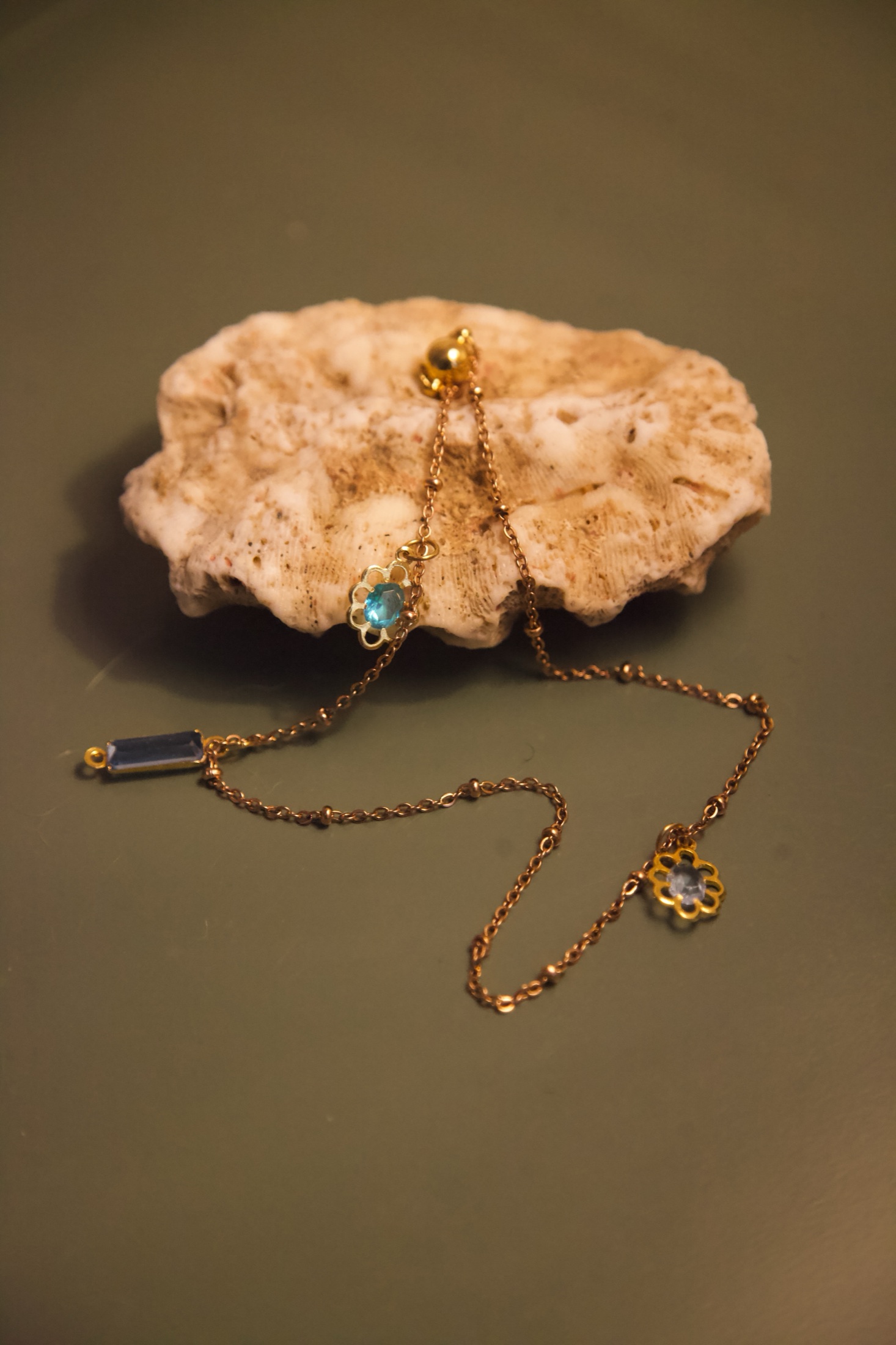 Gold charm jewellery arranged across a pale coral stone on a deep green surface.
